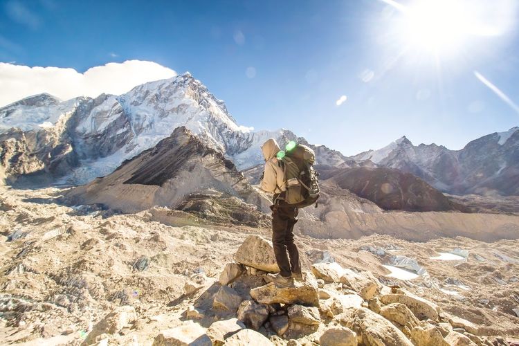 Side view of backpacker standing on rock by mountains against sky during sunny day
