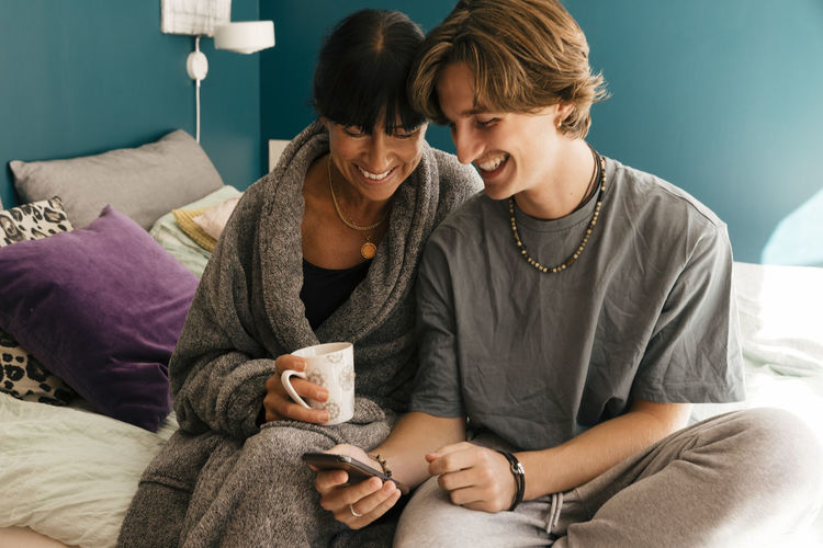 Happy mother and son sharing smart phone while sitting on bed in bedroom at home
