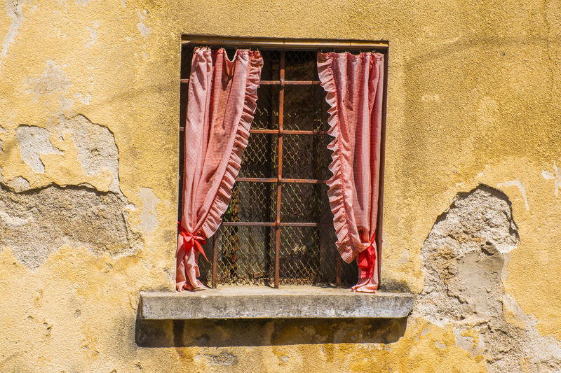 Window with rusty railing and red curtains on old yellow wall peeling from time