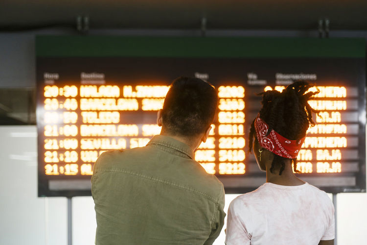 Young man and woman discussing over arrival departure board at railroad station