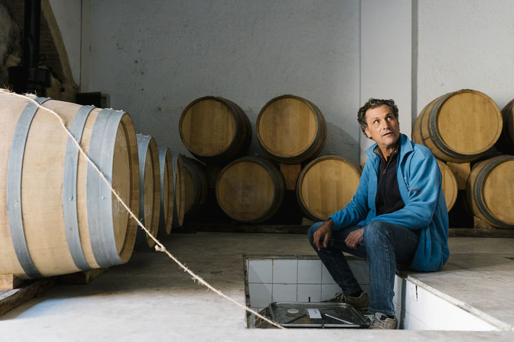 Mature male worker sitting amidst barrels at winery