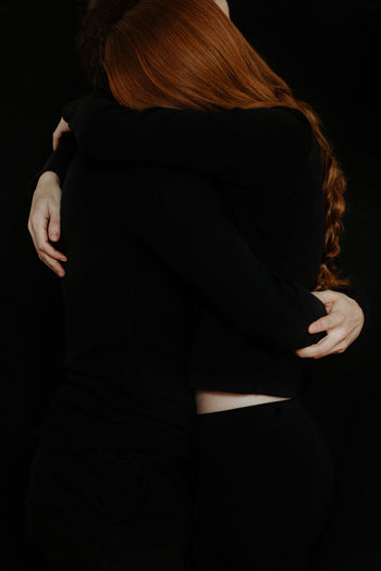 Unrecognizable side view of cropped tender boyfriend tenderly embracing redhead girlfriend while standing in dark studio on black background