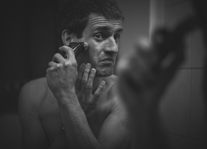 Portrait of shirtless young man holding electric razor reflected in the mirror