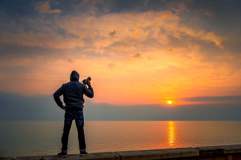 Rear view of man holding camera standing by sea against sky