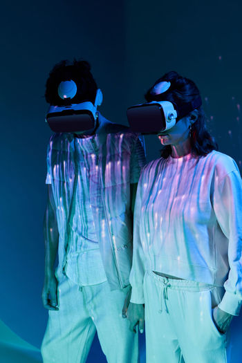 Couple in white wear and modern vr goggles standing in murk studio with glowing lights while exploring virtual reality together