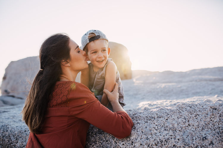 Mother whispering while smiling boy sitting on rock against clear sky
