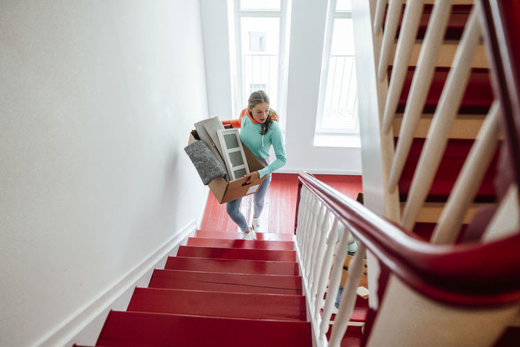 Young woman carrying cardboard boxes while moving up on staircase at home
