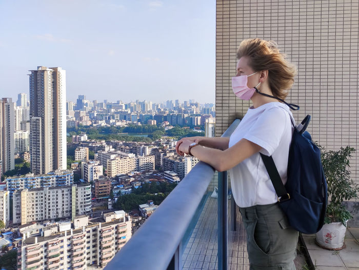 Woman blond woman in a medical mask with a backpack staying on the roof looking at the city