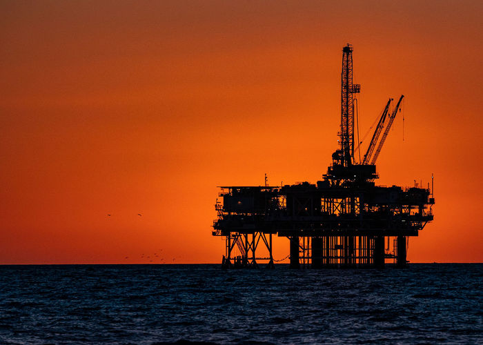 Silhouette oil rig in sea against orange sky