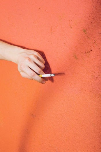 Cropped image of hand holding cigarette on orange wall
