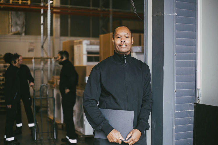 Confident male entrepreneur standing with clipboard at logistics warehouse