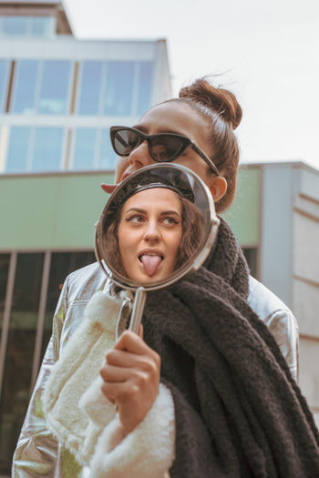 Woman sticking out tongue while holding mirror in city