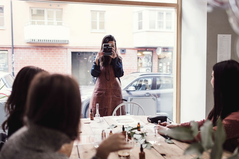 Woman standing against window photographing colleagues sitting at table in workshop