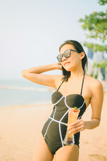 Midsection of young woman holding sunglasses at beach against sky