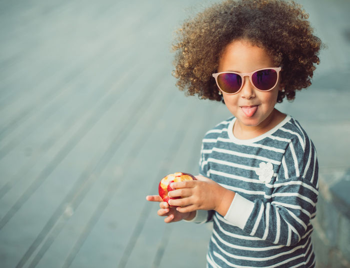 Cute little ethnic girl with afro hair wearing stylish striped shirt and sunglasses smiling and showing tongue while standing on city street and eating apple