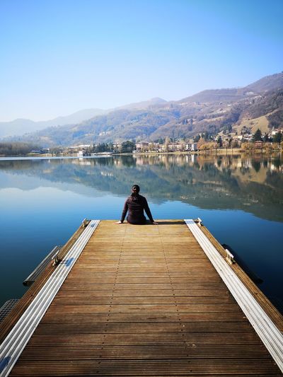 Rear view of woman sitting on pier over lake against mountains
