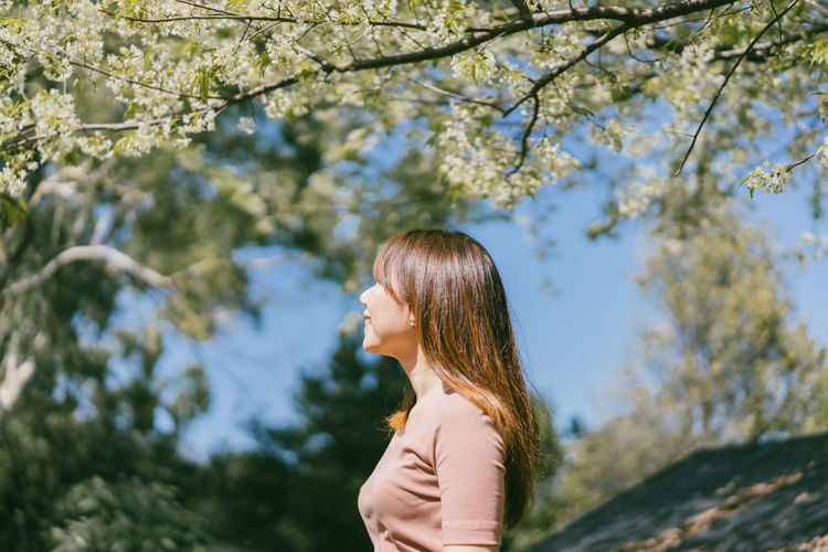 Side view of smiling woman standing below tree braches during sunny day