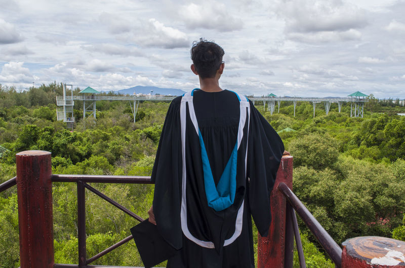 Rear view of man wearing graduation gown at observation point against forest