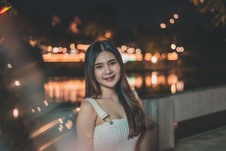 Portrait of smiling young woman standing on bridge over river at night