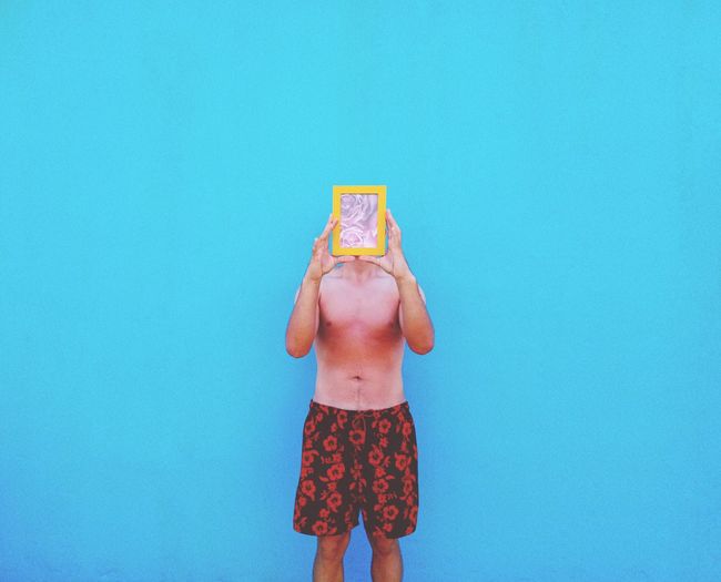 Shirtless man holding picture frame in front of face against blue wall
