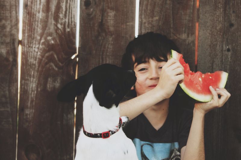 Smiling holding watermelon while sitting with dog against wooden fence