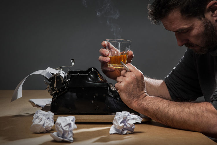 Man with drink and cigarette sitting by typewriter against black background