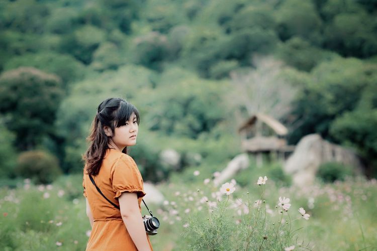 Beautiful young woman carrying camera while standing amidst plants on field