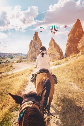 Rear view of woman riding horse against hot air balloons flying over landscape