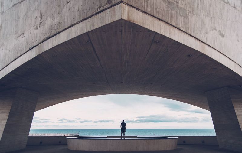 Rear view of man standing under bridge by sea