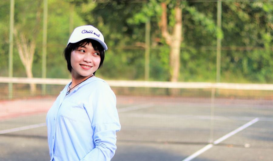 Beautiful asian woman with short hair, wearing hat and smiling broadly on tennis court