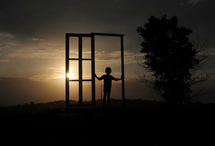 Silhouette boy standing at gate on field against sky during sunset