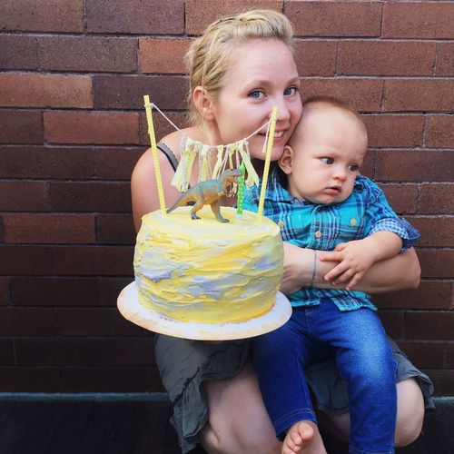 Portrait of smiling mother with cute son holding cake while crouching against brick wall