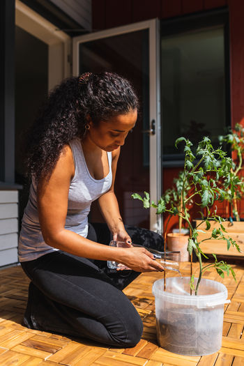 Young african american female on sunlit terrace of apartment clothes watering indoor plant with pure water