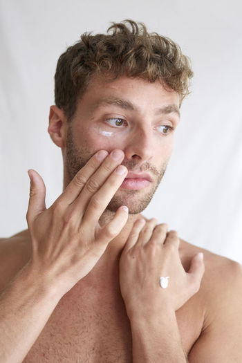 Shirtless young man applying cream on face against wall