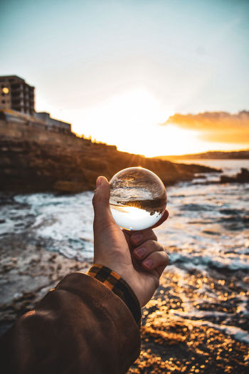 Cropped hand holding crystal ball against sea during sunset