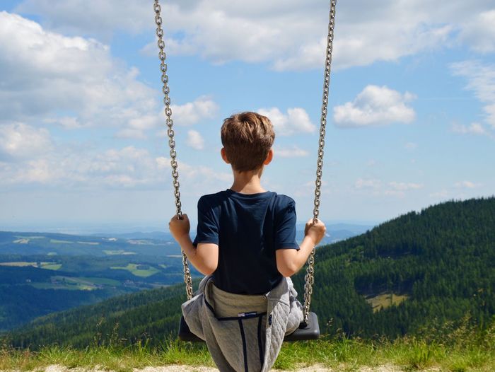 Rear view of boy swinging by landscape against cloudy sky