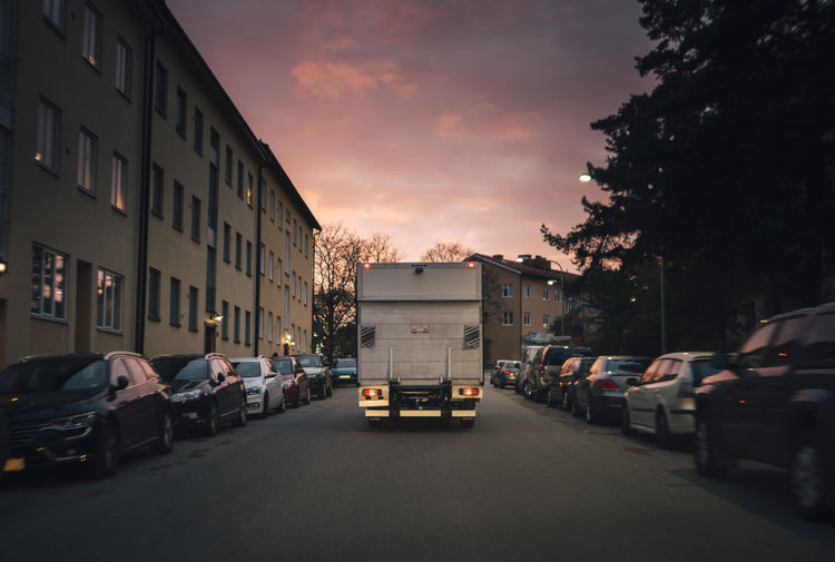 Delivery van moving on road amidst parked cars in city against sky at sunset