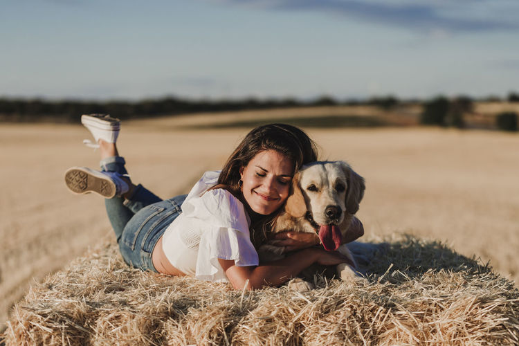 Happy mid adult woman with dog lying on hay at agricultural field