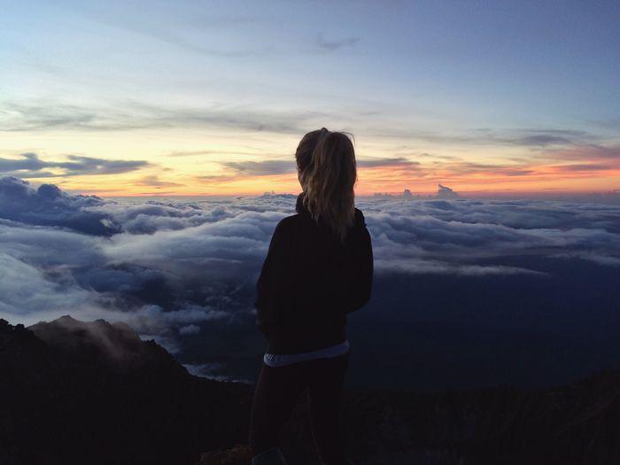 Rear view of woman standing on top of mountain over clouds during sunset