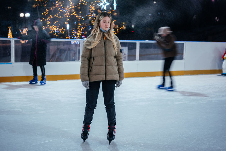 A blonde teenage girl skating on the skating rink with christmas lights on the background