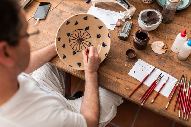 From above of crop faceless man sitting at table with brushes and drawing sketches on handmade ceramic plate