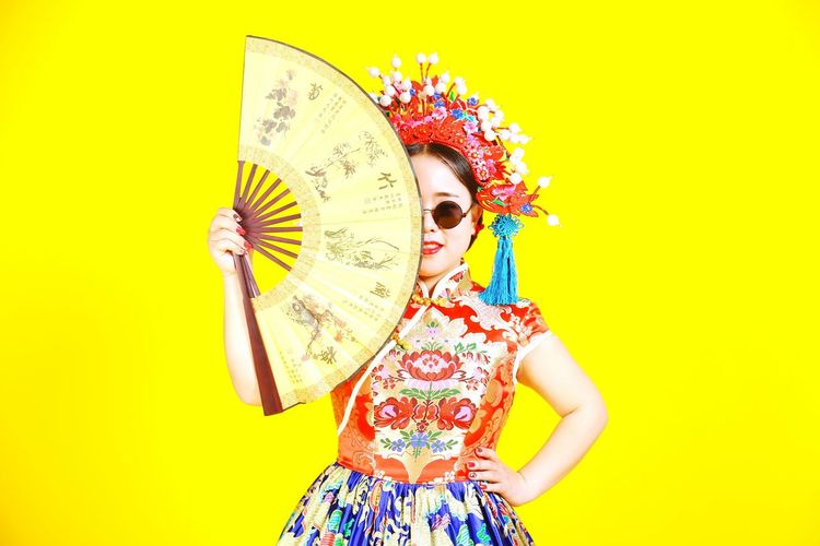 Woman in traditional clothing holding hand fan while standing against yellow background