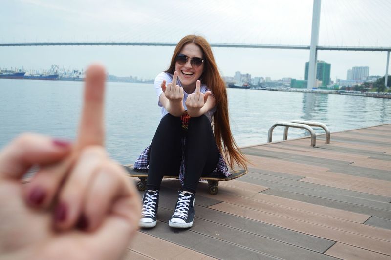 Laughing young woman shows middle fingers to camera while sitting on a skateboard near the sea