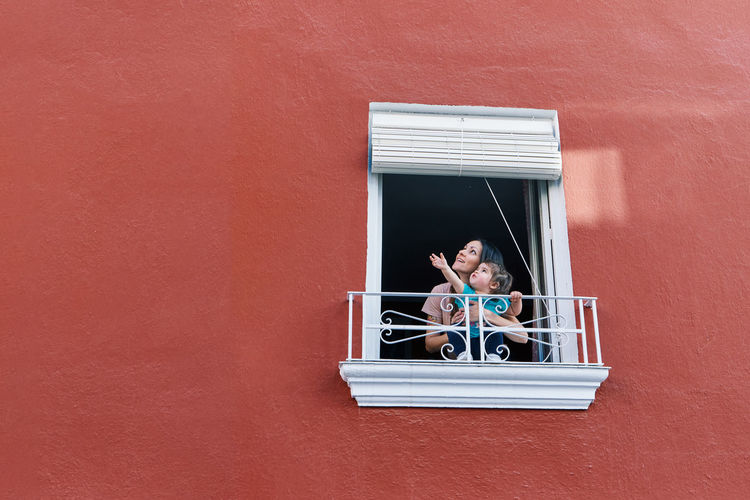 Woman clapping in the window of her house with her daughter on a red background