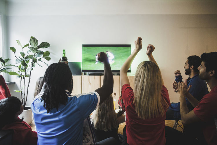 Cheerful friends with arms raised watching soccer match in living room