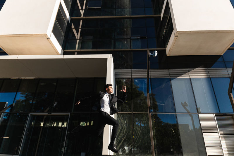 From below of male entrepreneur in formal suit in moment of jumping above ground on background of modern glass building showing concept of rush