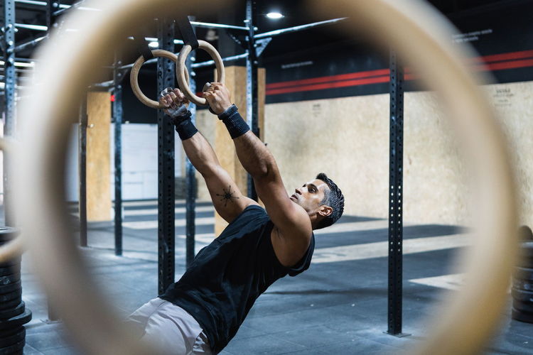 Side view of through blurred sportive equipment of side view strong hispanic male athlete doing exercise with gymnastic rings during training in gym