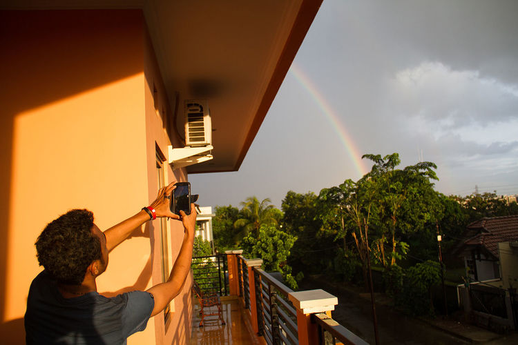 Rear view of man photographing rainbow in cloudy sky