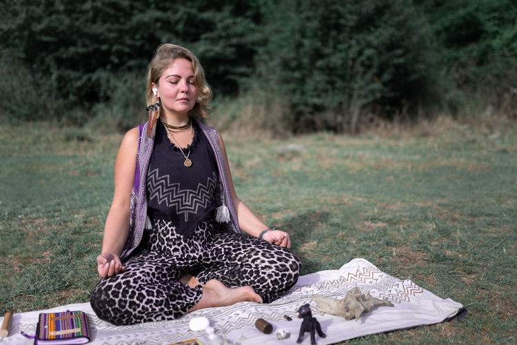 Female sitting in lotus pose on blanket near healing crystals with wireless earphones listening to music and meditating with closed eyes in nature