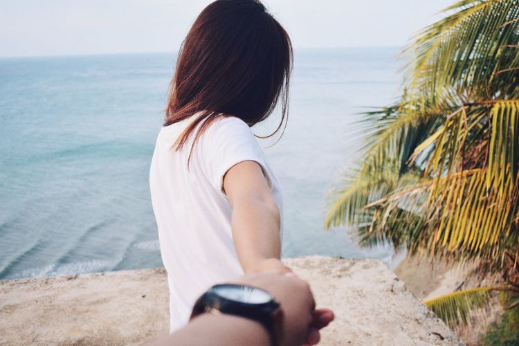 Cropped image of man holding woman hand on retaining wall by sea